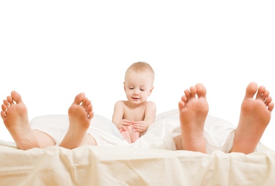 Bebê sentado na cama ao lado dos pais - Foto: maga/ShutterStock