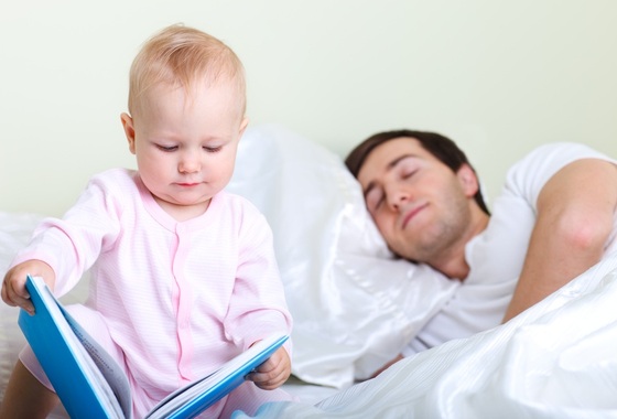 Bebê com livro na mão e papai dormindo na cama - Foto: BlueOrange Studio / ShutterStock