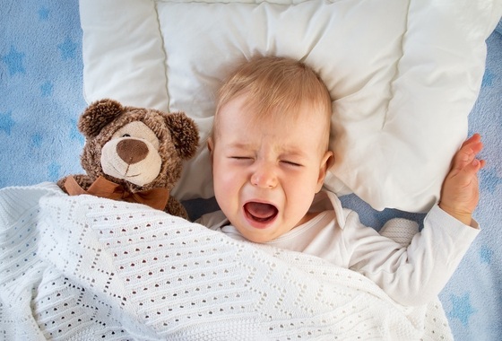 Bebê chorando deitado na cama ao lado de seu ursinho - foto: Anna Grigorjeva/ShutterStock.com