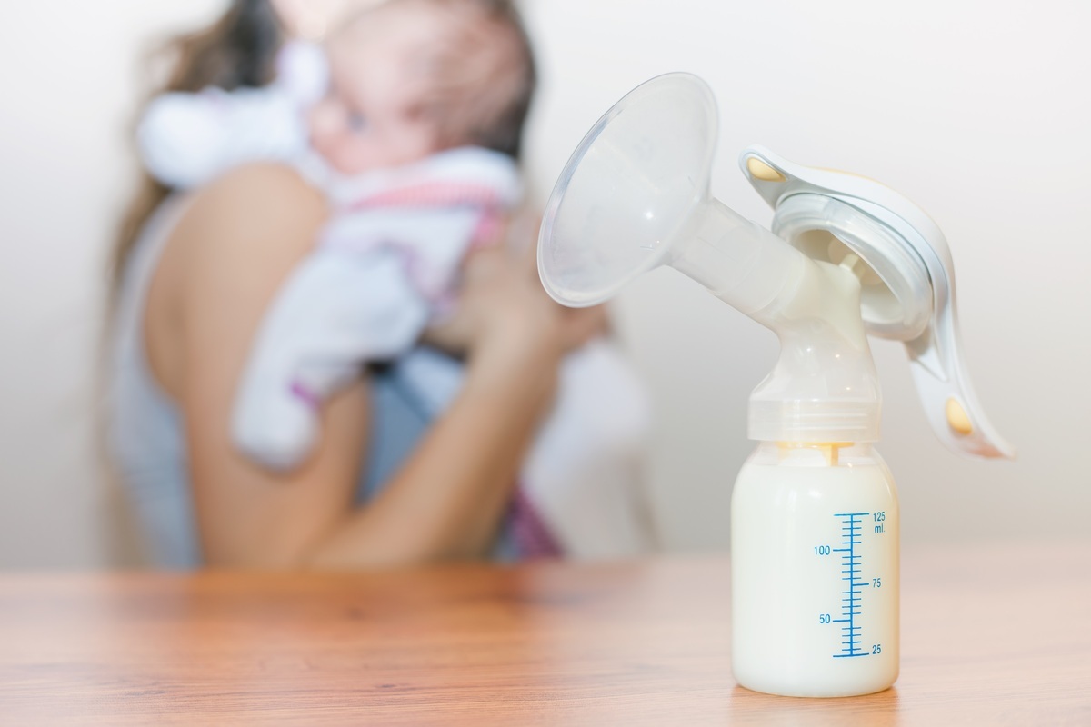 Bomba de extração de leite materno e mãe com o bebê no colo ao fundo - foto: Pavel Ilyukhin/ShutterStock.com